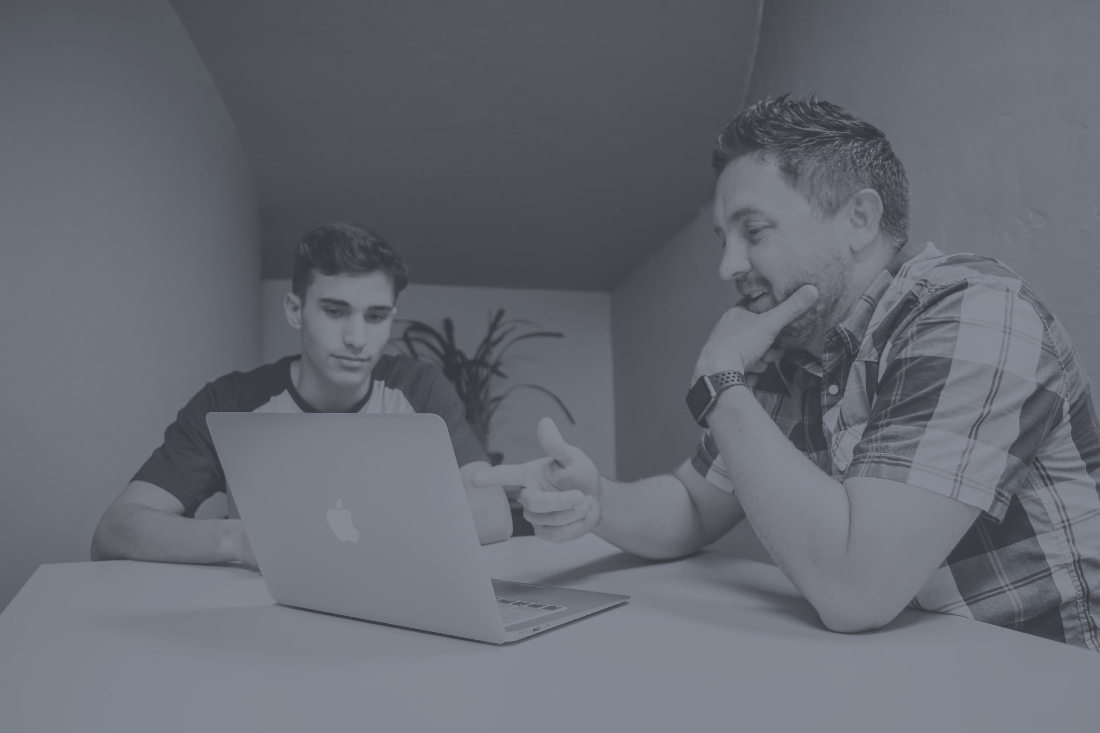 Two men sit looking at a laptop, discussing it thoughtfully 